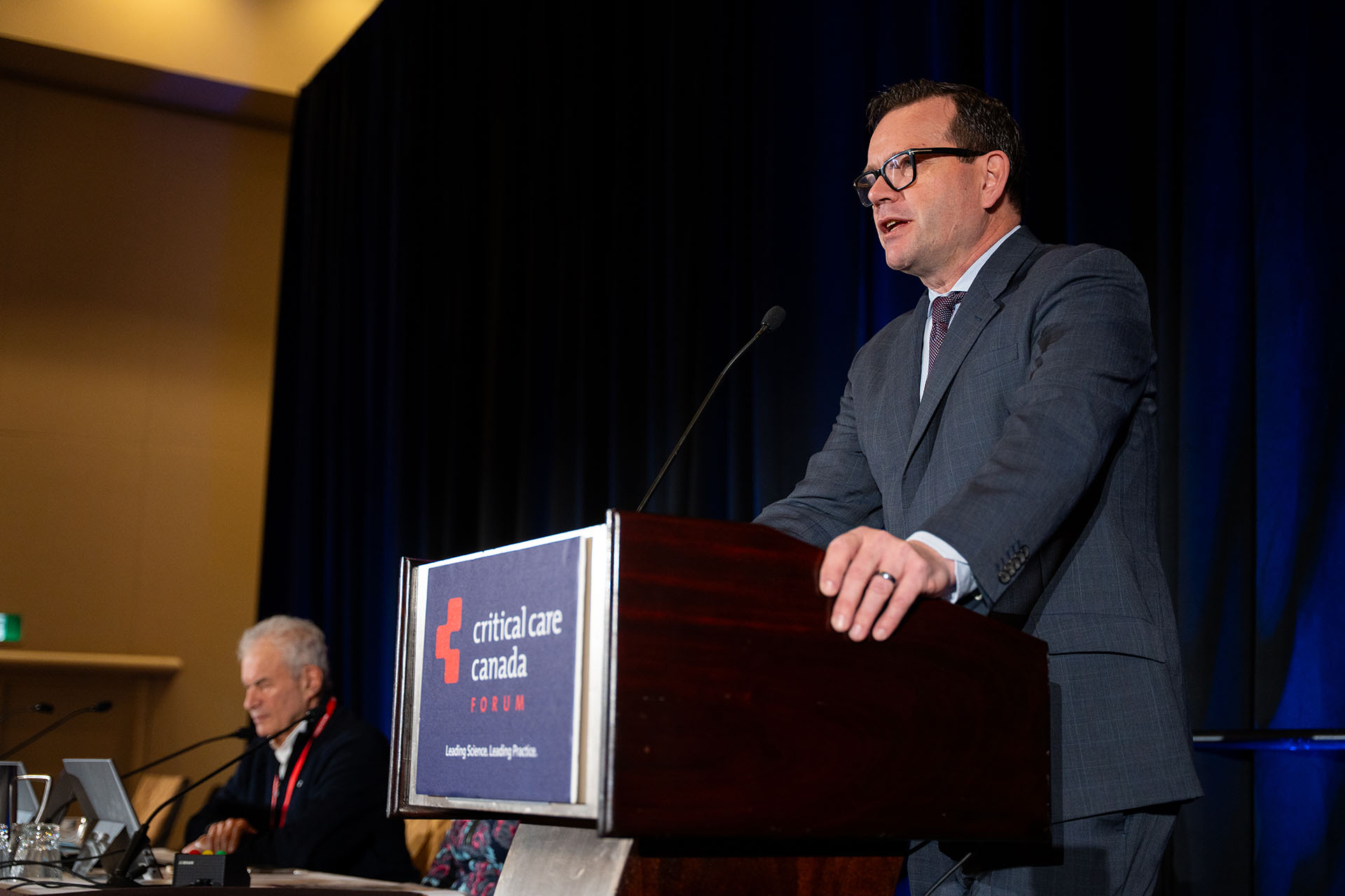 Man speaking at wooden podium with microphone and blue sign at conference with dark blue curtain