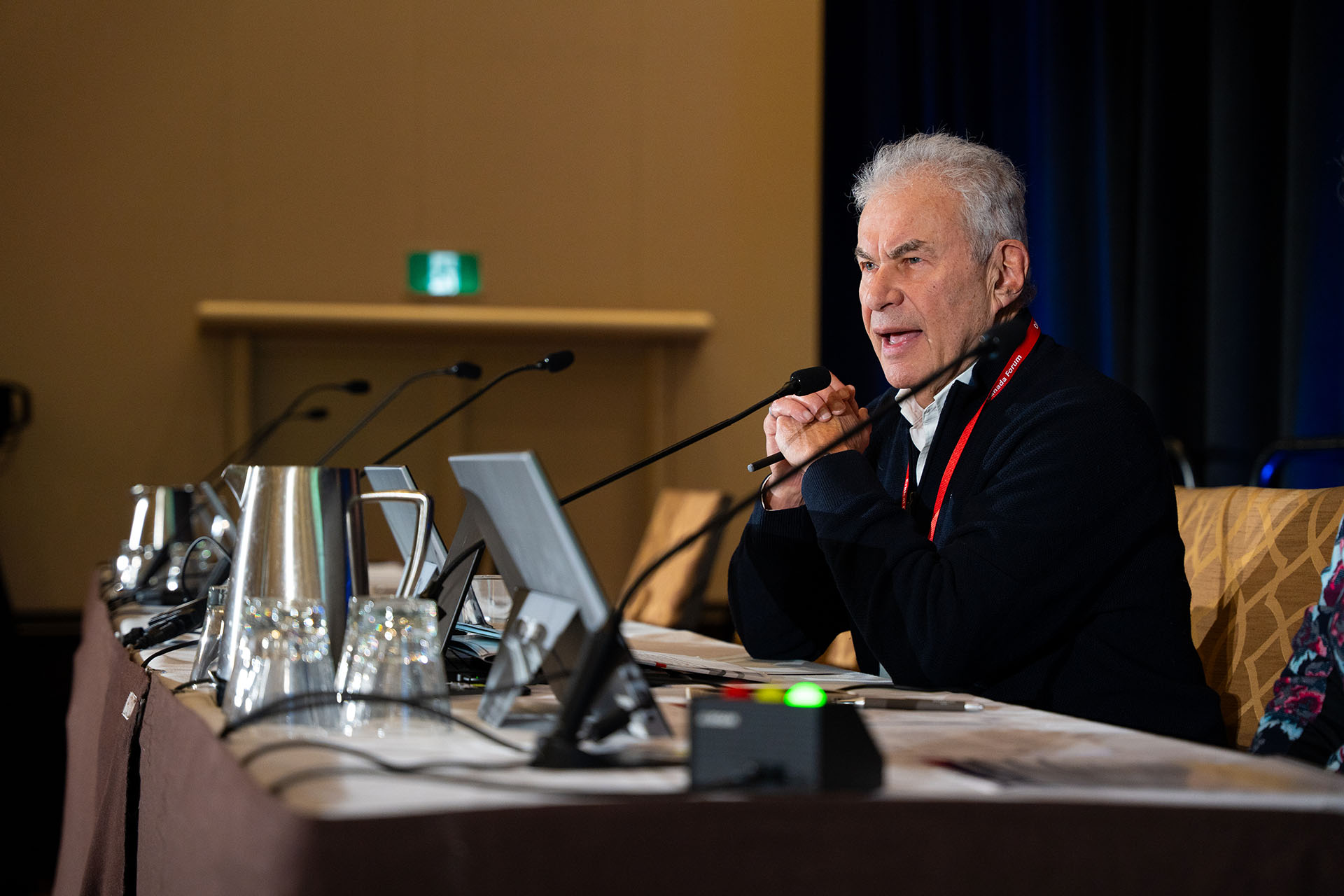 Man speaking at conference table with metal water pitchers, glass cups, and electronic devices, brown and silver