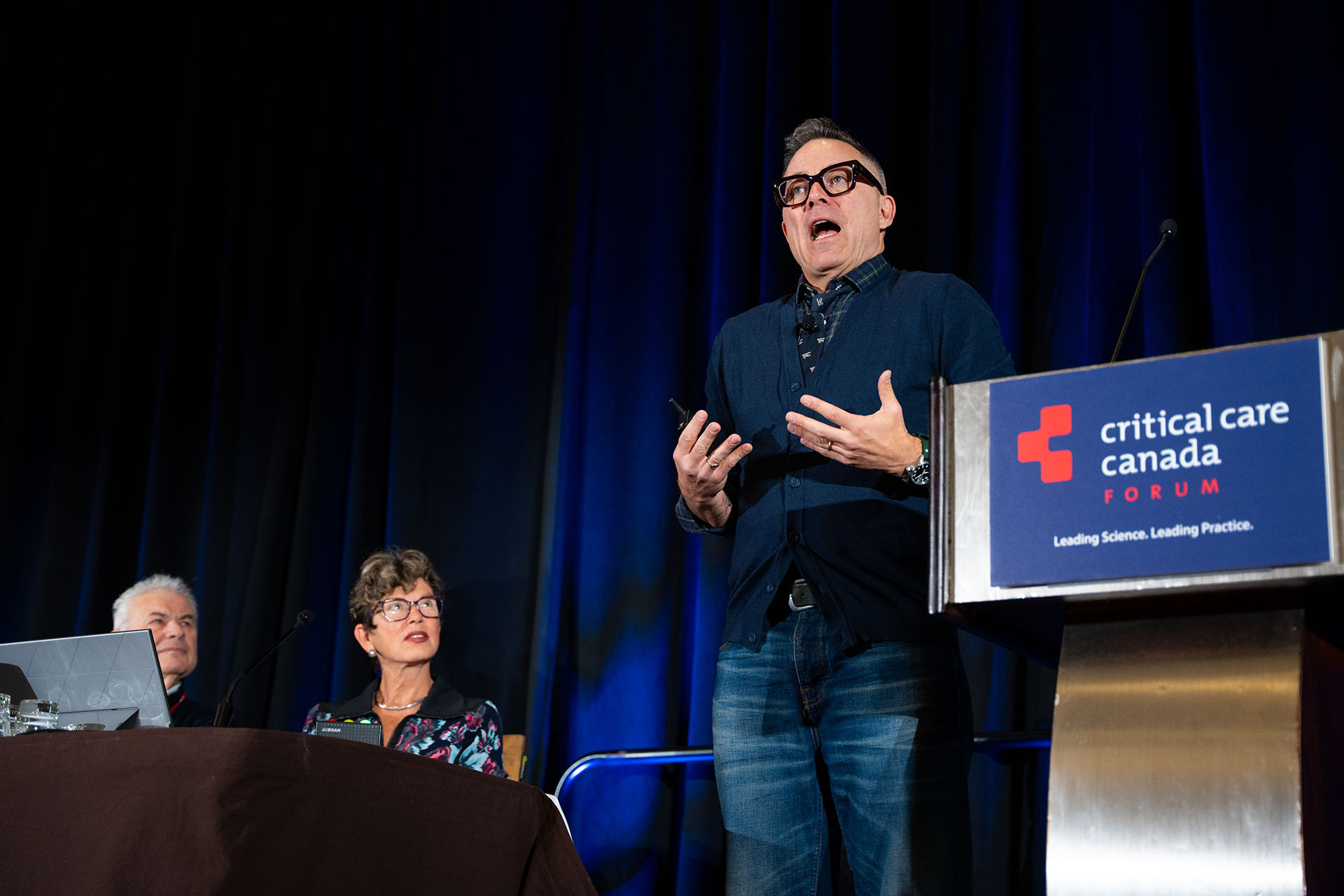 Man speaking at podium with blue fabric sign, two seated people at table with dark cloth, dark blue curtain background