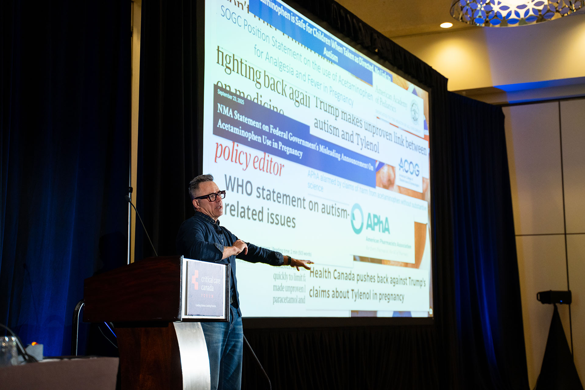 Man presenting at podium with wooden lectern and large white projection screen with text in conference room