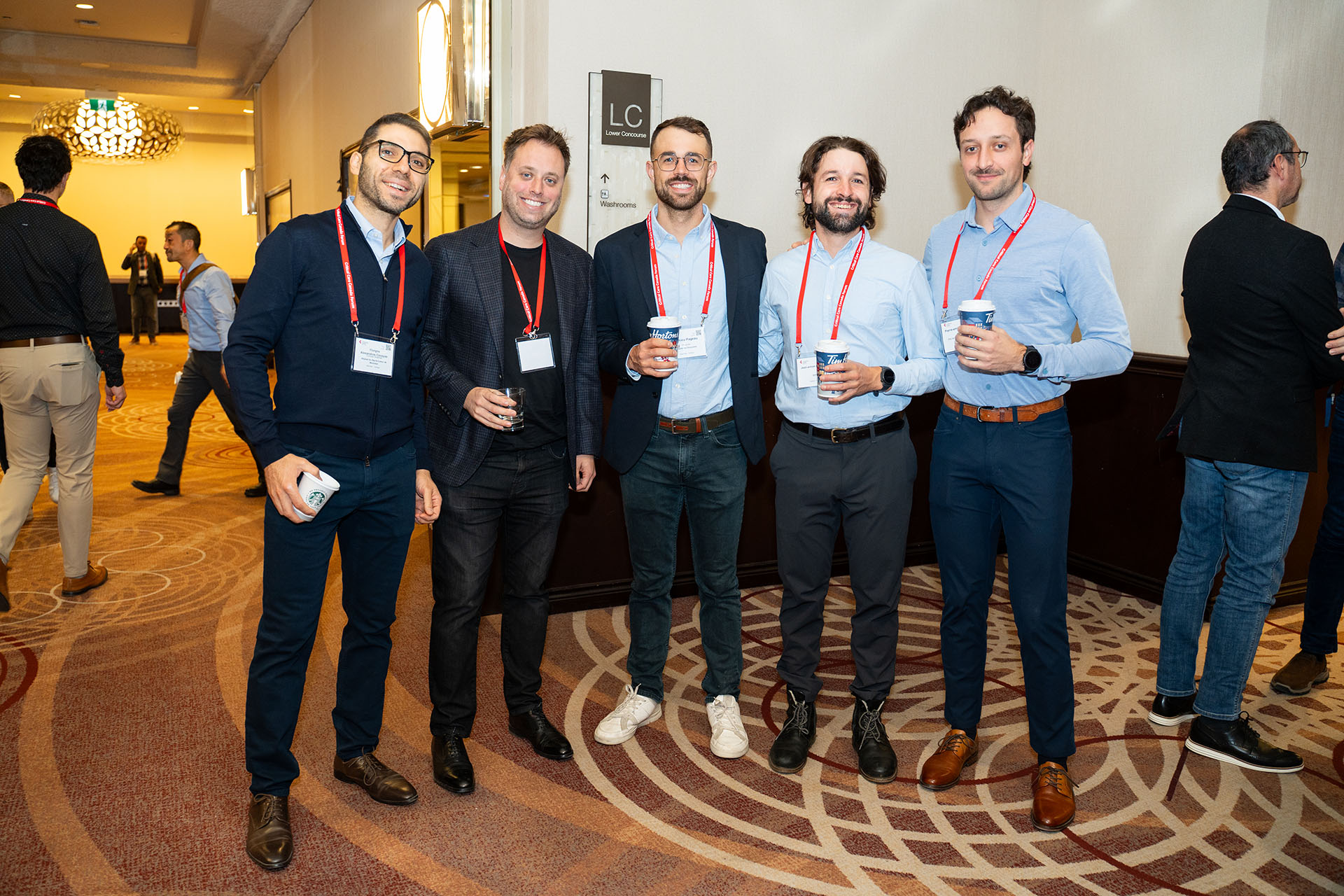 Group of five men wearing business casual clothes and red lanyards holding paper coffee cups indoors
