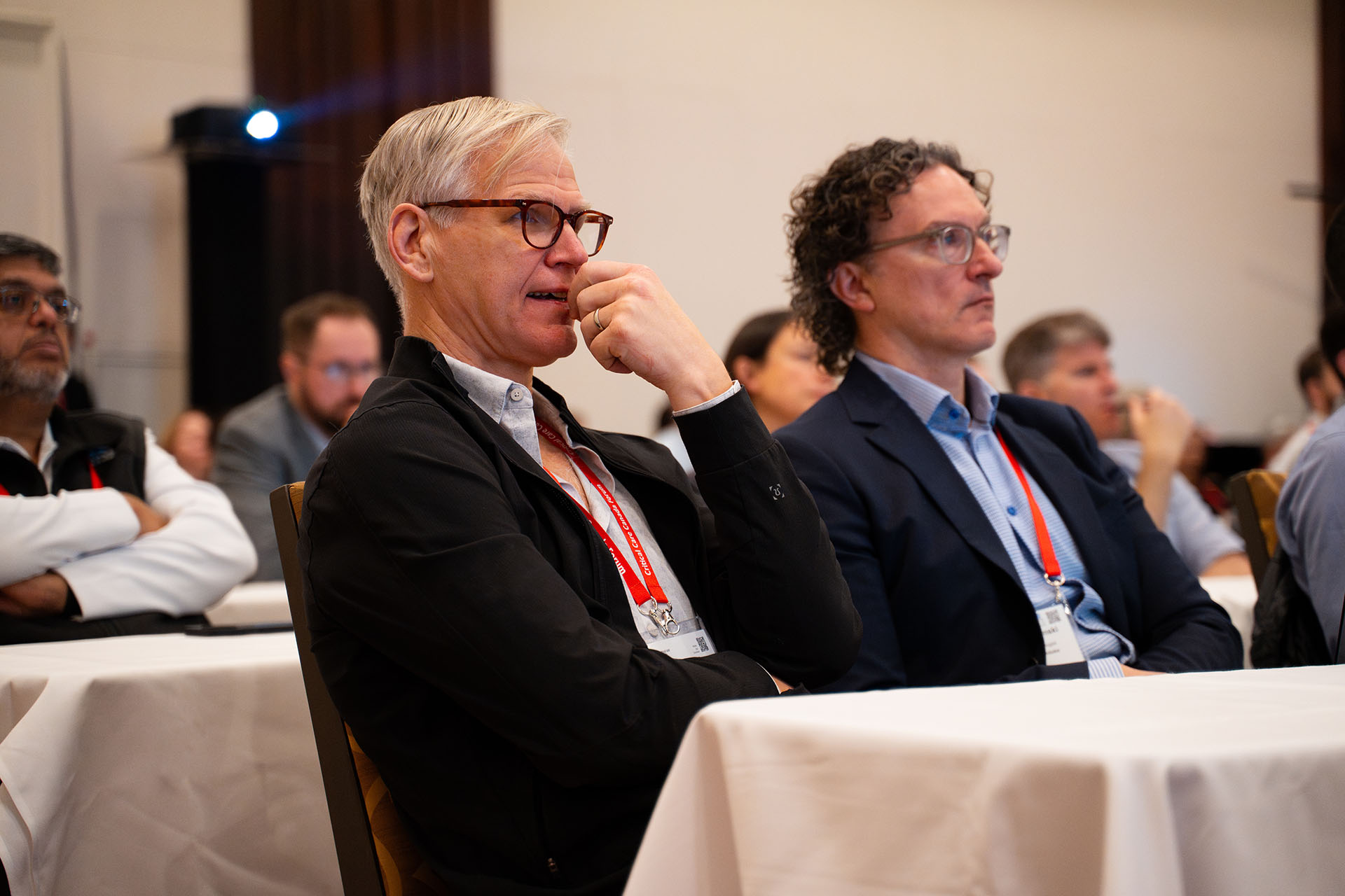 Men sitting on wooden chairs wearing glasses and lanyards at a conference with white tablecloths