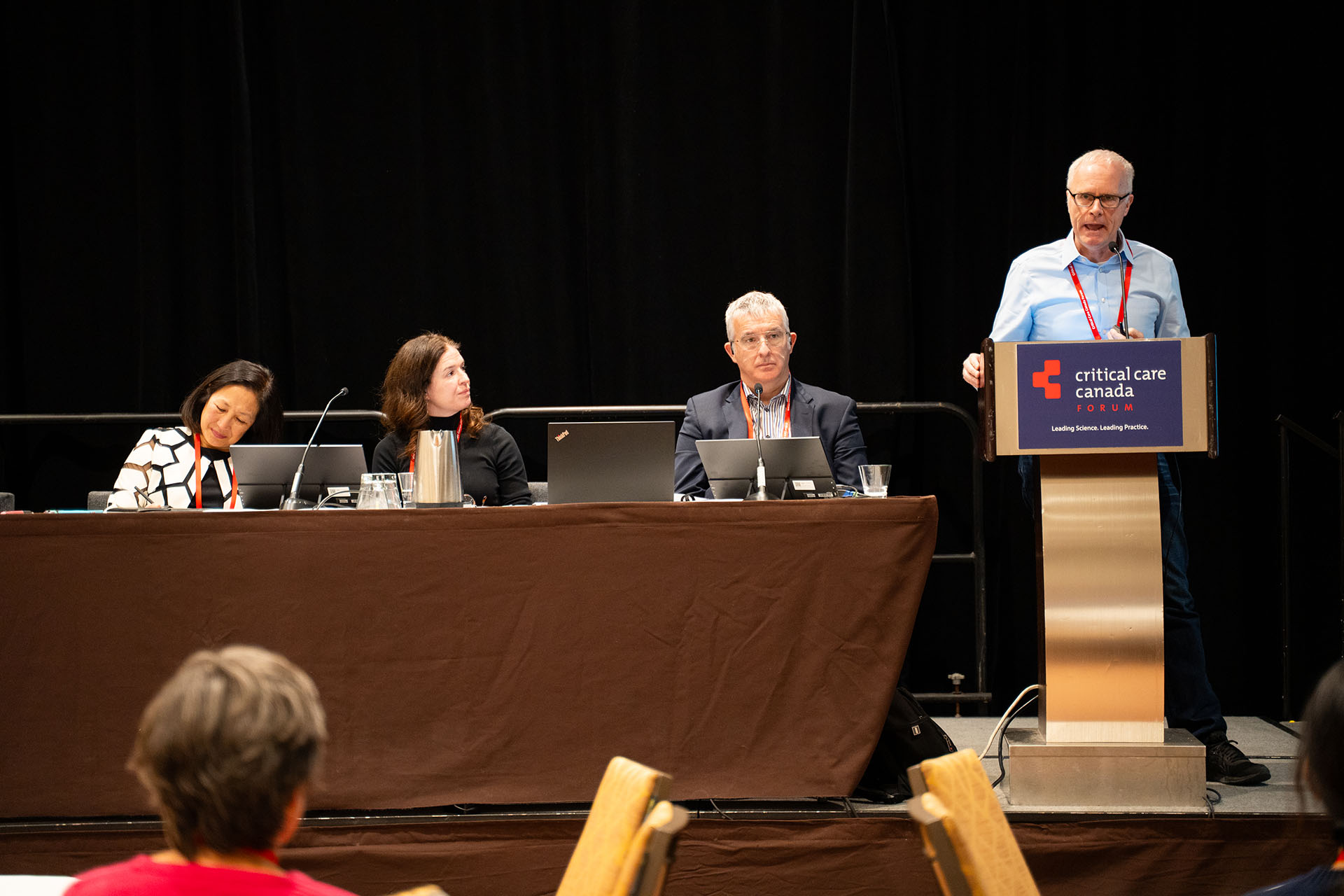 Conference panel with four people, laptops, metal pitcher, and brown fabric-covered table in a dim room