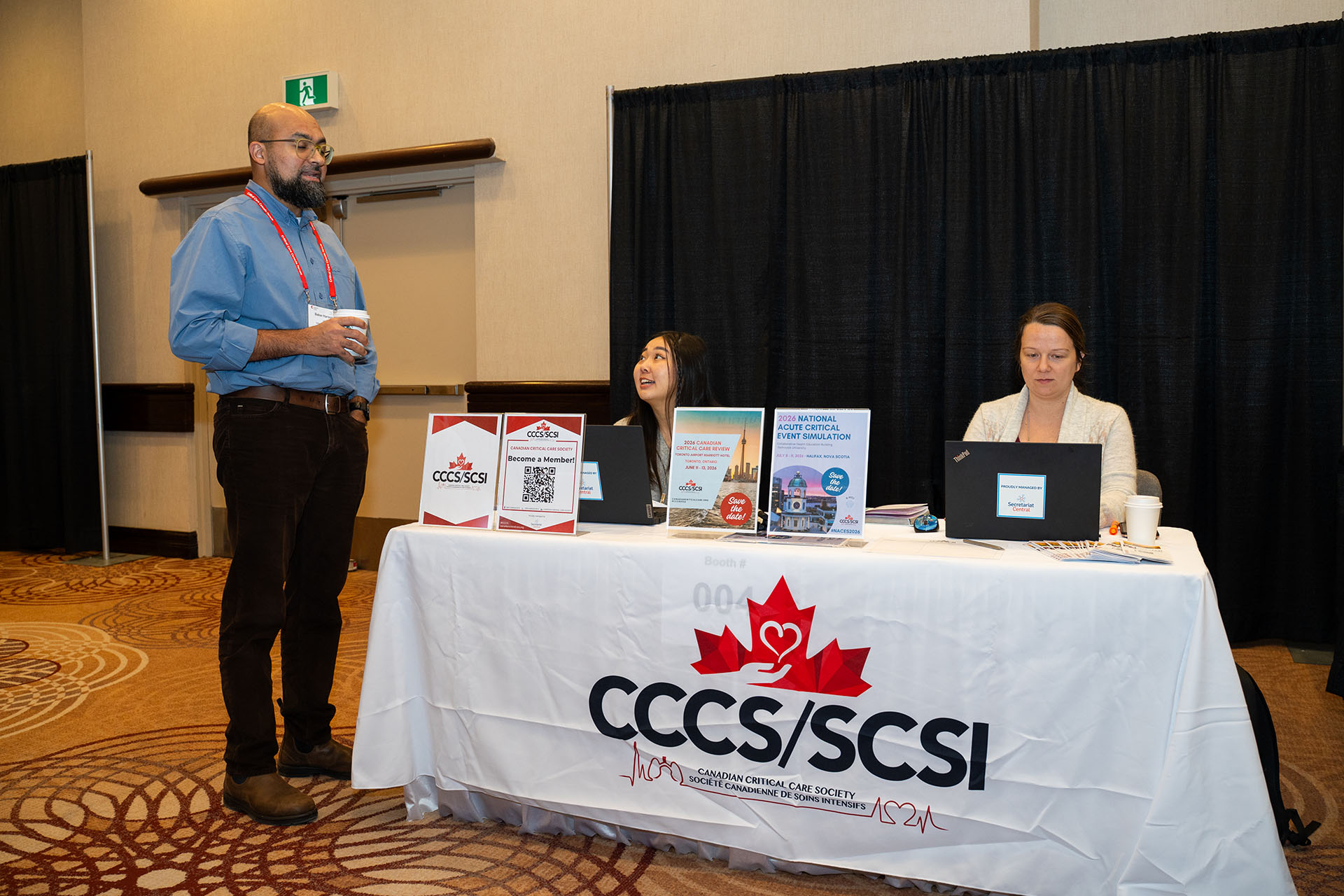 Event booth with white fabric tablecloth, promotional signs, two laptops, and three people interacting indoors