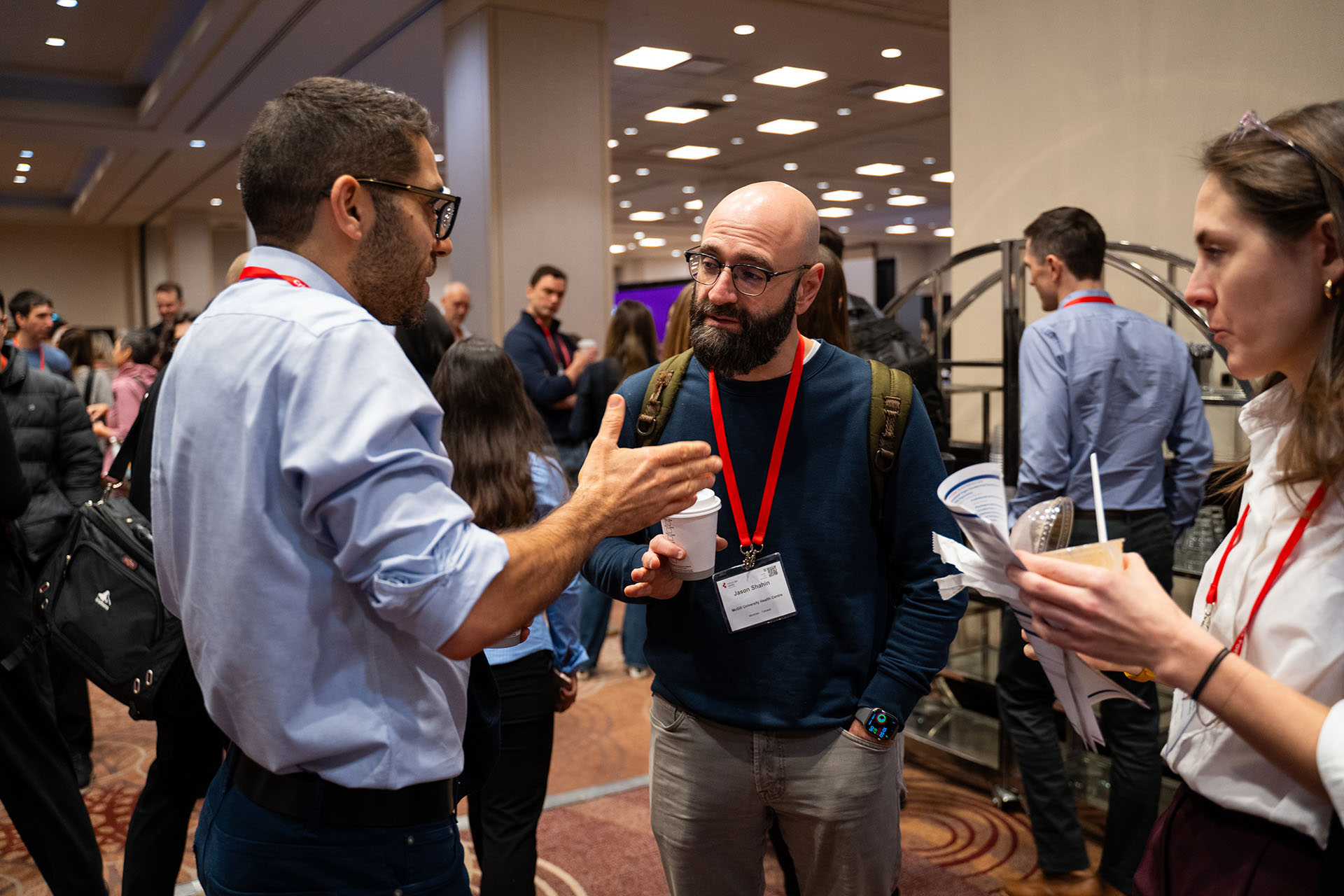 People wearing fabric clothing and lanyards talking and holding paper cups in a carpeted conference room