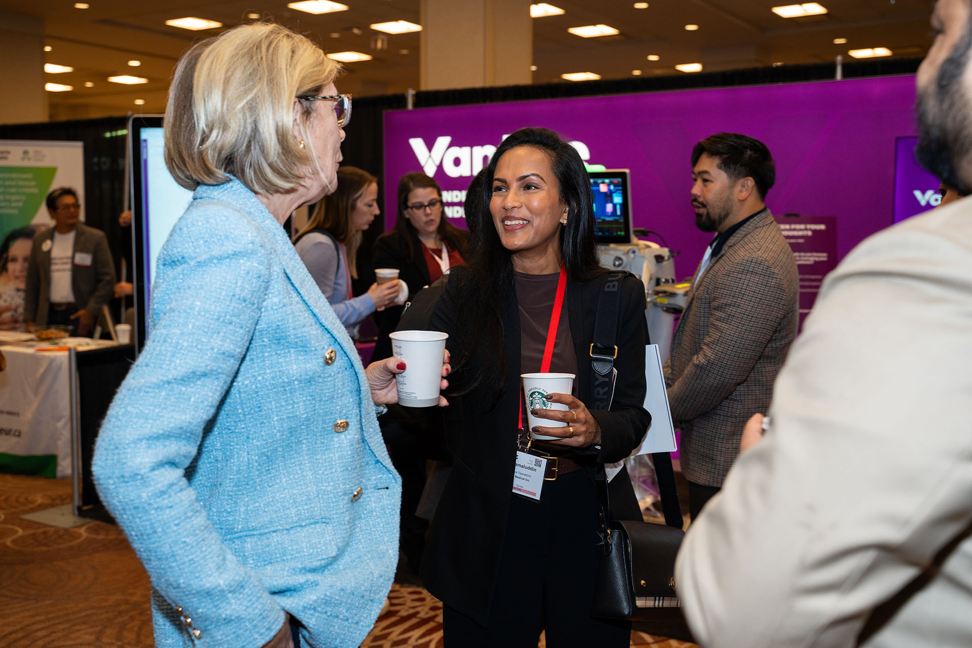 People holding paper cups and talking at an indoor event with fabric clothing and a purple backdrop
