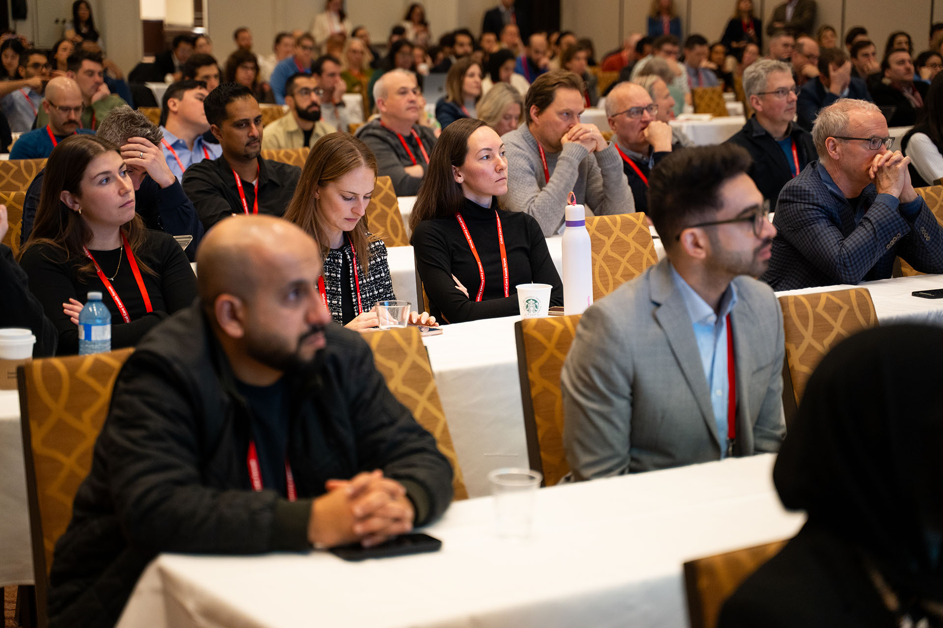 Audience of people sitting on patterned fabric chairs at a conference with white tablecloths