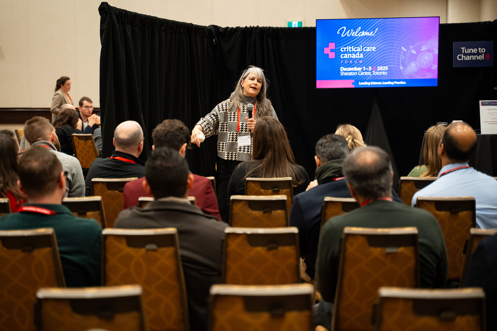 Speaker holding microphone wearing patterned wool sweater addressing seated audience on wooden chairs