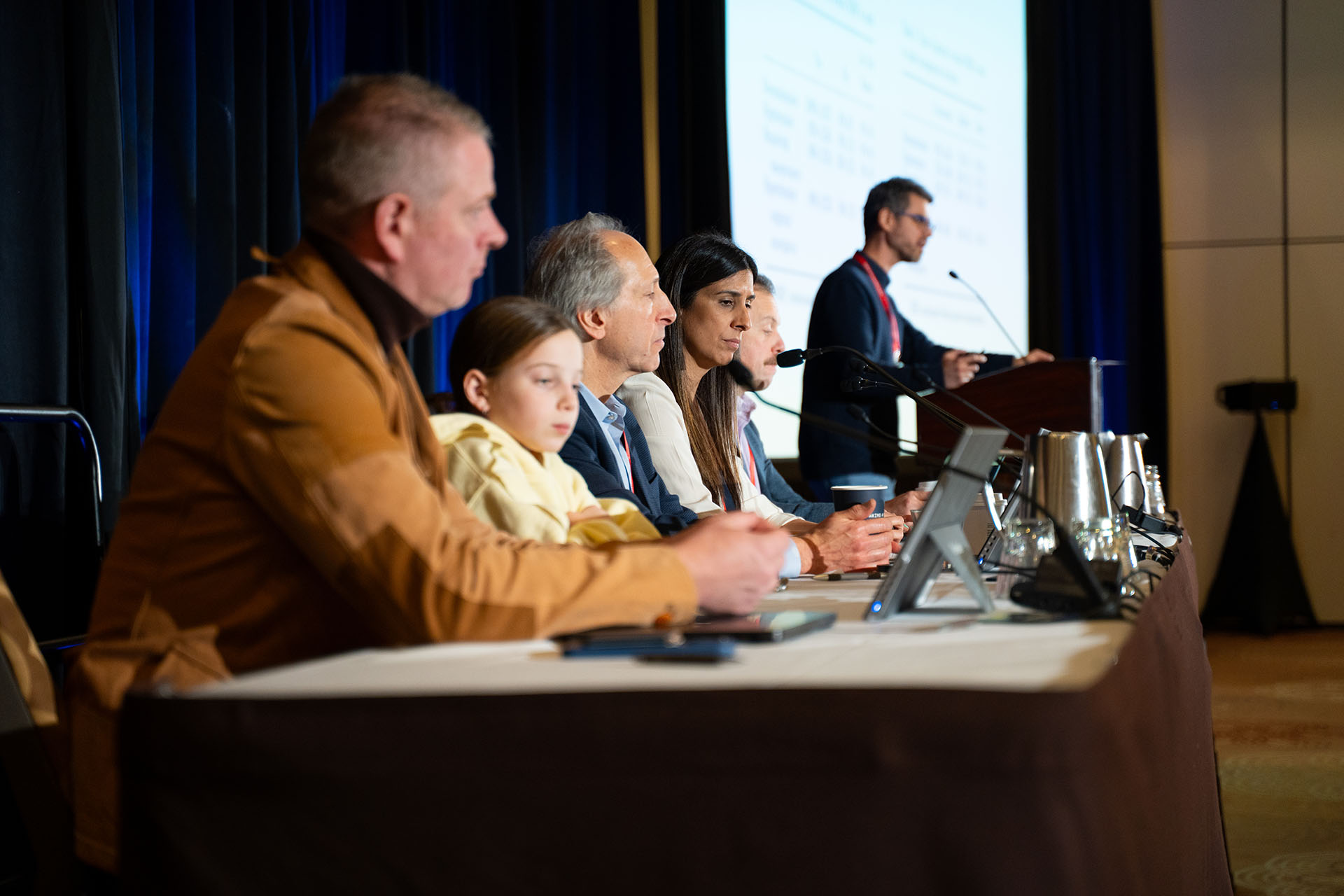 Panel of people sitting at a table with microphones, laptops, and metal water pitchers in a conference room